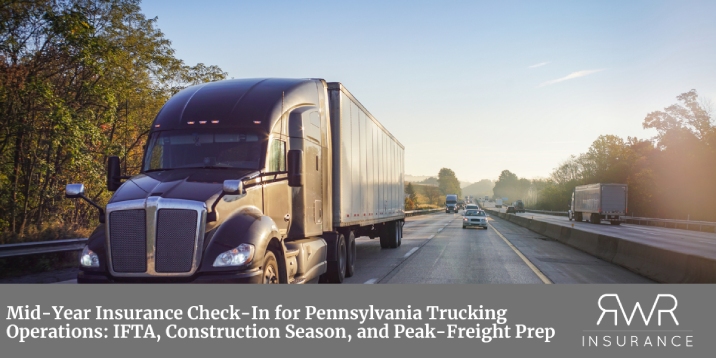 A large, dark brown semi-truck drives on a multi-lane highway under a clear, bright sky, with other vehicles and dense green trees visible in the distance. The bottom portion of the image features a solid grey banner containing white text that reads, "Mid-Year Insurance Check-In for Pennsylvania Trucking Operations: IFTA, Construction Season, and Peak-Freight Prep." In the bottom right corner of the banner is a stylized white logo for "RWR INSURANCE." - www.rwrtruckinginsurance.com